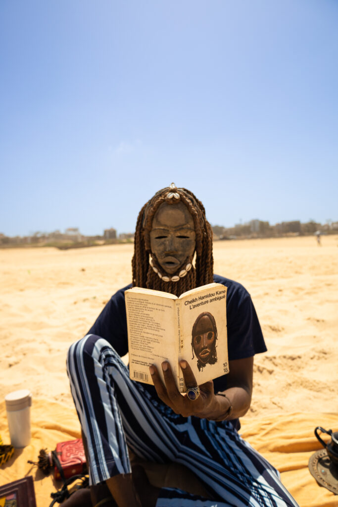 A figure seated in a desert reading a book, wearing an embellished top and blue wrap