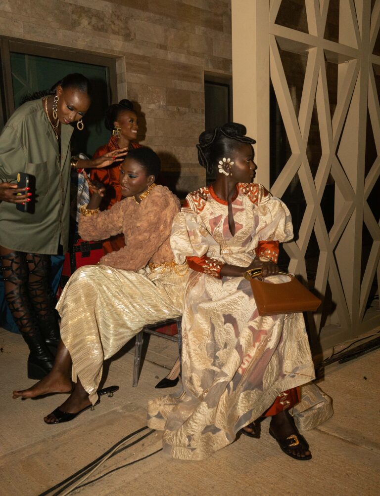 Three African fashion models backstage at Dakar Fashion Week, featuring one standing in an olive green dress and two seated wearing elegant gold and bronze patterned ensembles.