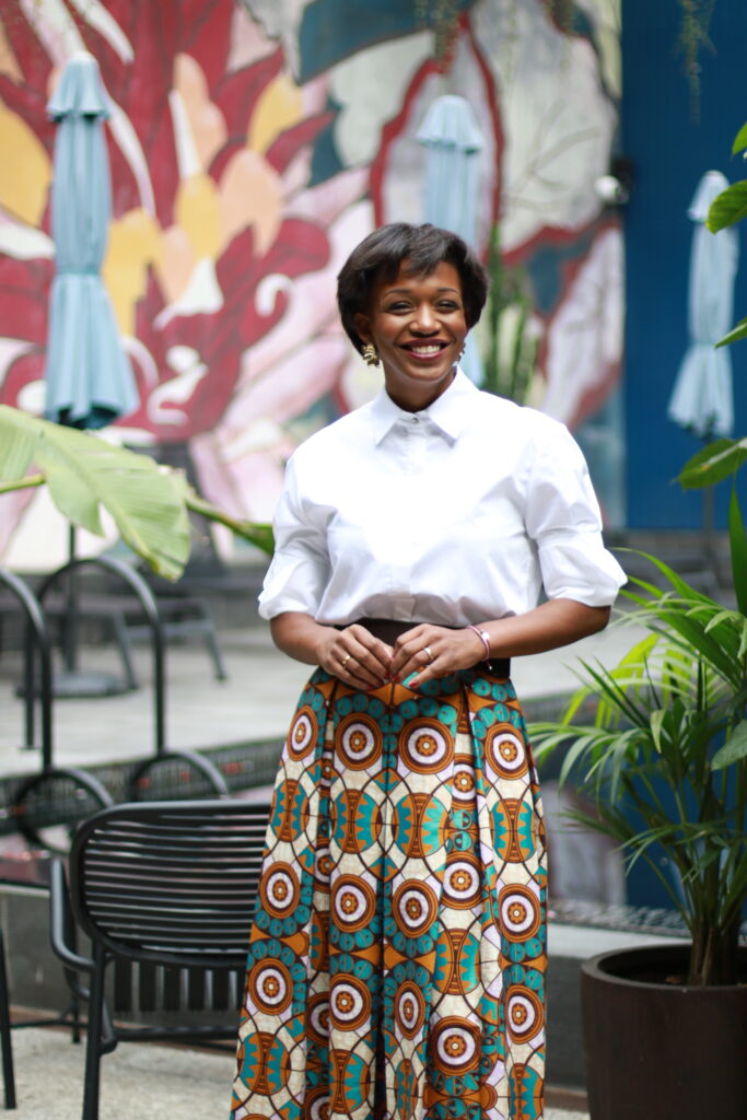 A Decade of Design: Roselyn Silva on Intentional Elegance and Her São Tomé Roots 2 Roselyn Silva in a white shirt and patterned skirt seated before a painted mural