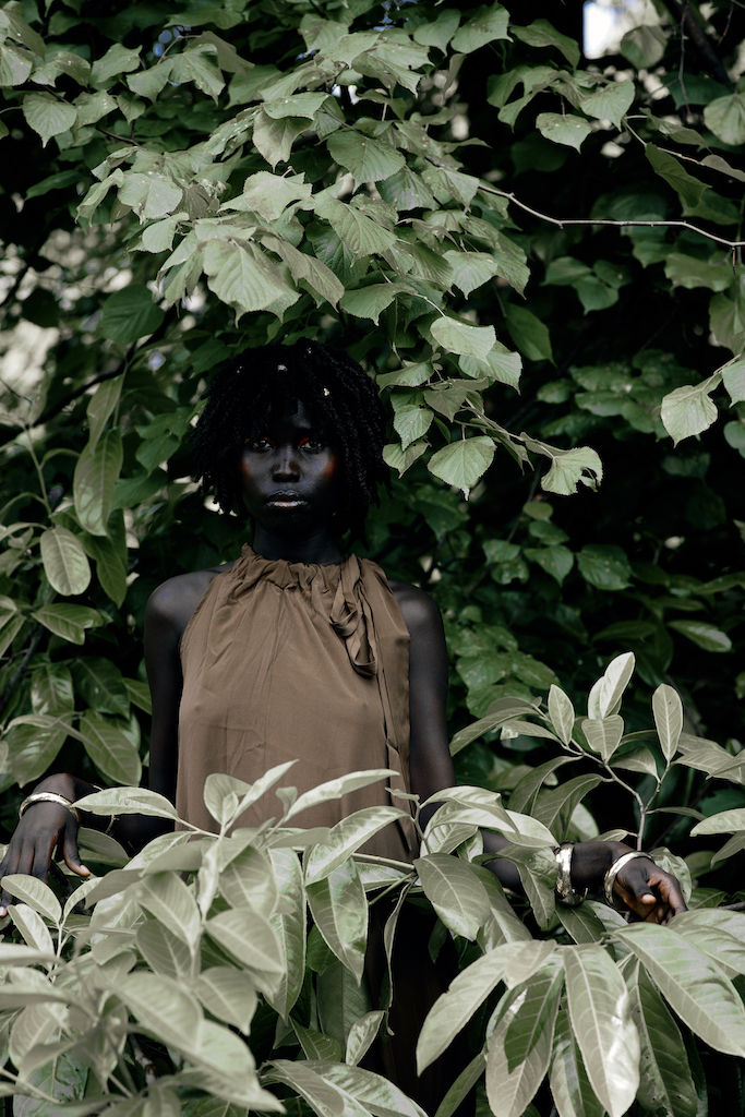 Portrait of a woman in a sleeveless brown dress standing amid dense green foliage.
