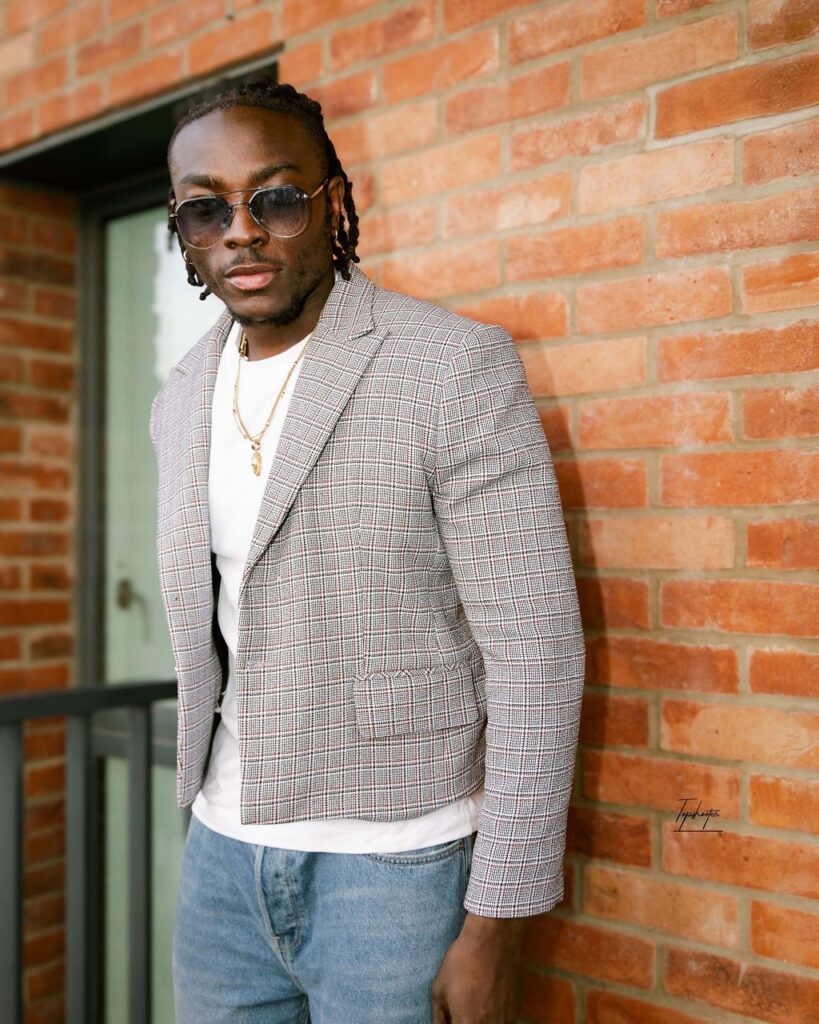 Stylist Sodiq Ayinde poses against a brick wall, wearing sunglasses, a checked blazer over a white T-shirt, light denim jeans, and layered gold chains.