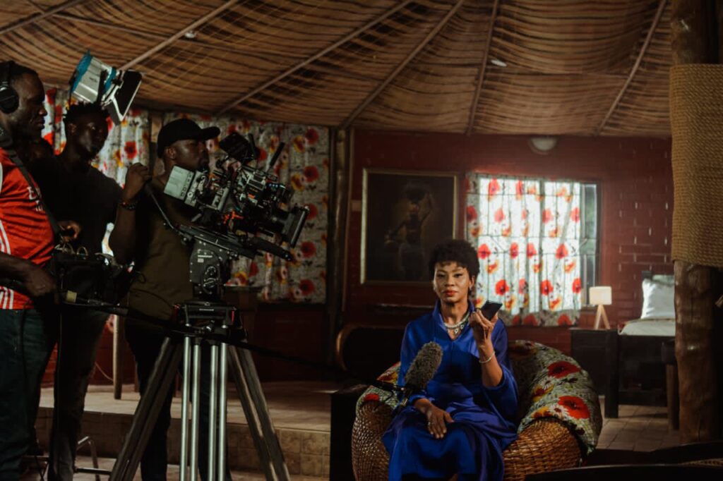 A behind-the-scenes shot of actress Nse Ikpe-Etim seated in a wicker chair on the set of A Devil Is A Liar. She wears a deep blue dress, holding a phone, with a film crew and camera setup surrounding her.