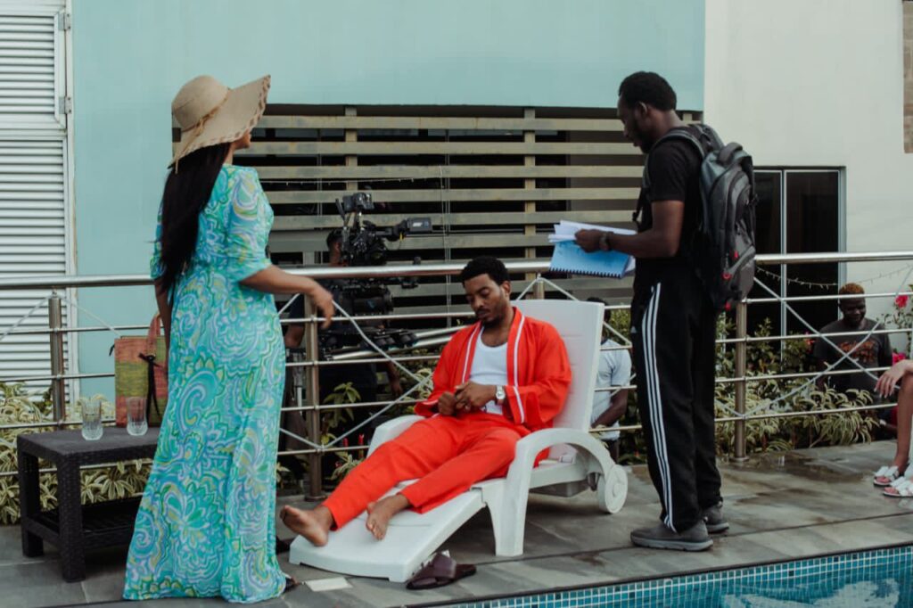 Behind-the-scenes image from the film A Devil Is A Liar. An actor lounges on a white pool chair in a bold red loungewear set styled by The Style Pundit, while crew members and a director engage nearby with scripts and cameras.