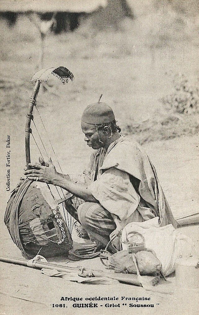 Early 20th-century griot in French West Africa, dressed in traditional attire and performing on a kora.