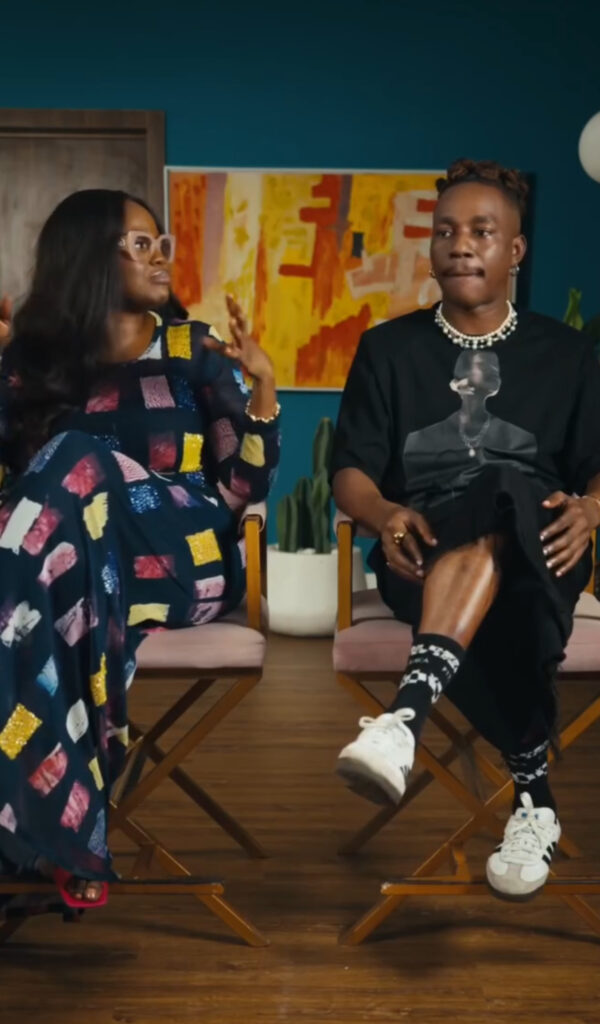 Dahmola and filmmaker Jade Osiberu seated side by side in conversation. Dahmola wears a black graphic T-shirt with pearl necklace and sneakers, while Jade Osiberu is dressed in a patterned long gown with colorful geometric patches. They are discussing the fashion direction for A Christmas in Lagos.