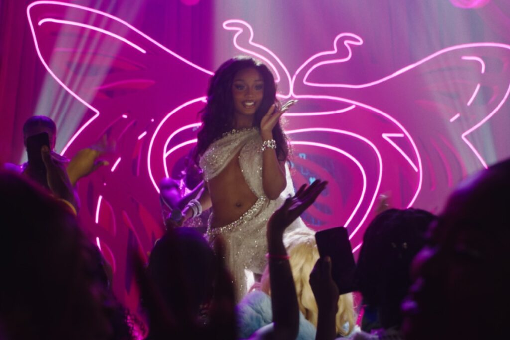 Ayra Starr performs on stage in a glittering draped outfit during a nightclub scene in A Christmas in Lagos. Neon pink butterfly lights glow behind her as the crowd cheers and films with their phones.