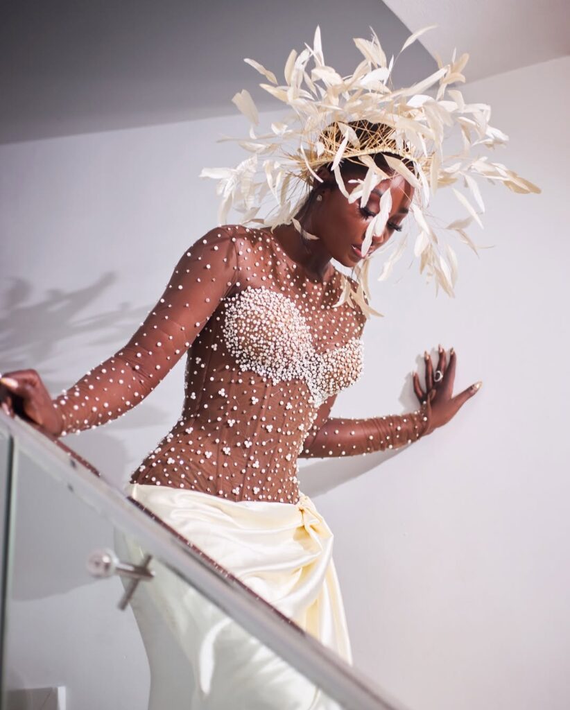 Tolu Bally descends a staircase wearing a sheer corset gown adorned with pearls, paired with a draped ivory skirt and a dramatic feathered headpiece.