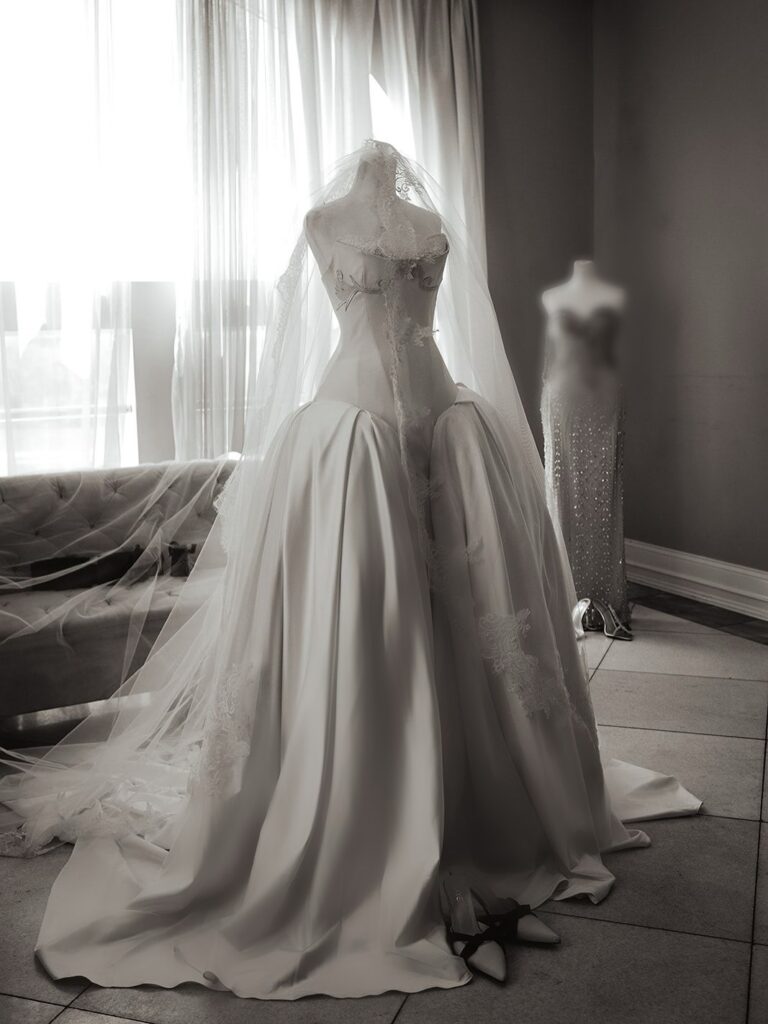 A headless mannequin displays a dramatic white bridal gown by Veekee James, featuring a cinched corset bodice, voluminous skirt, and lace veil, photographed in soft natural light.