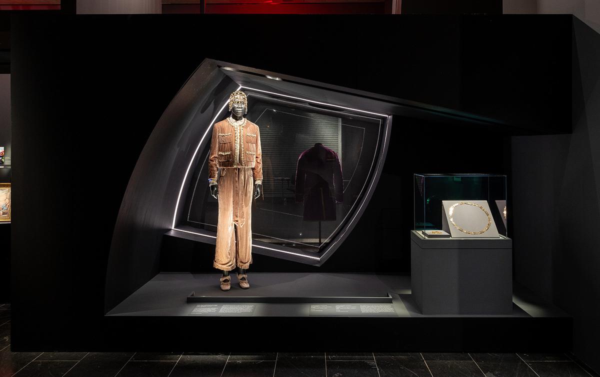 A mannequin in a beige, ornate jumpsuit with headpiece in a futuristic black display. Nearby, a gold necklace is showcased under soft lighting.