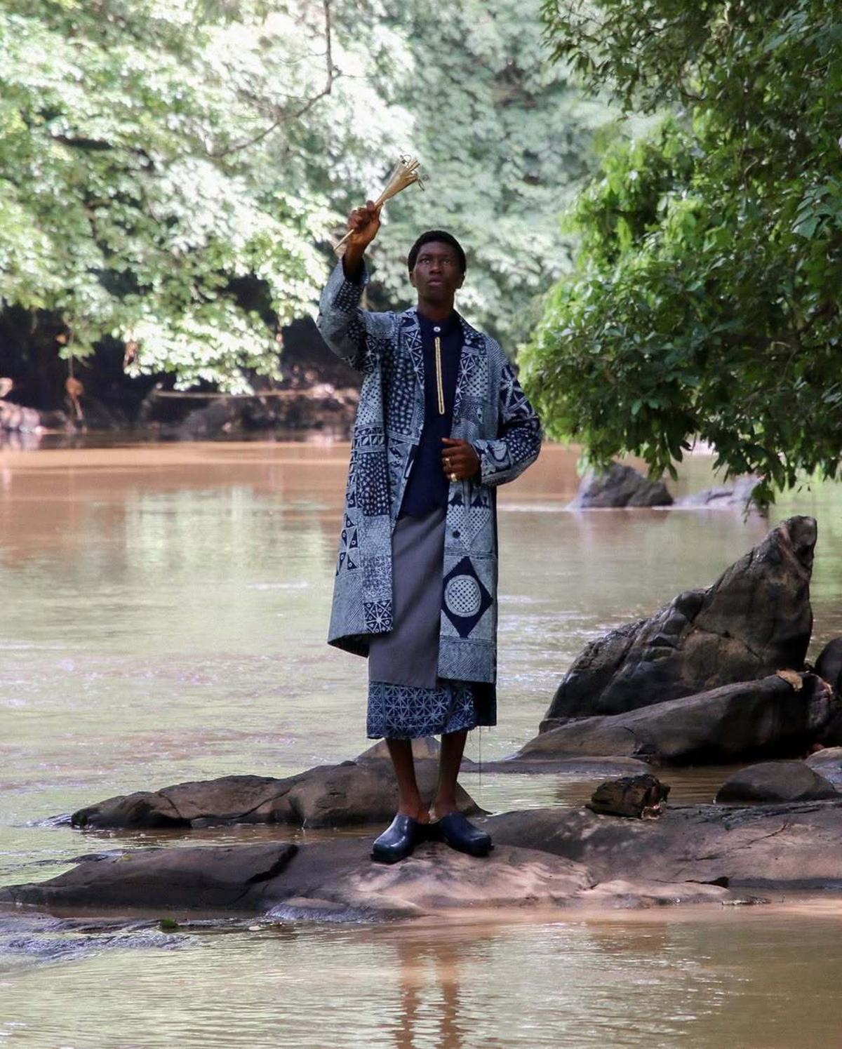 A person stands on rocks in a serene river, raising a small object. They wear a patterned coat, skirt, and shoes. Lush greenery surrounds them, conveying tranquility.