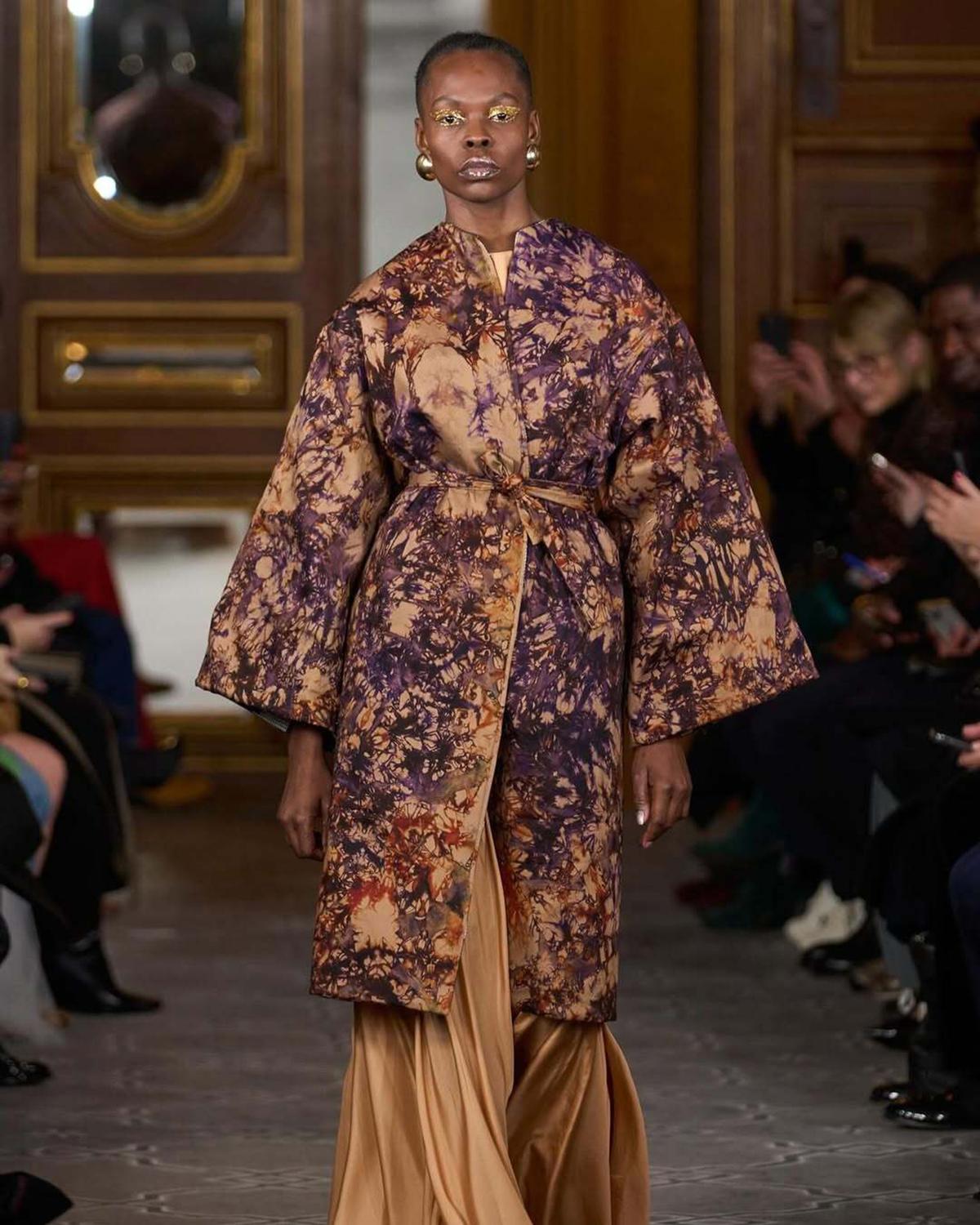 Model walks runway in an elegant, patterned coat with warm earthy tones, paired with flowing golden trousers. Audience watches in a grand room.