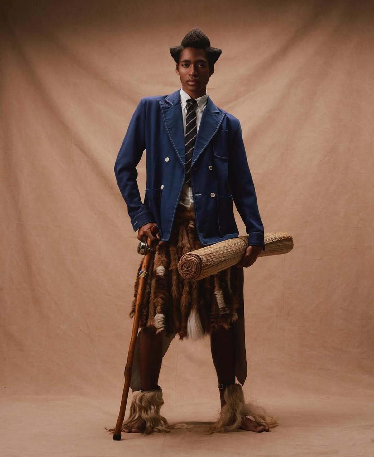 A person in a blue blazer with a tie stands confidently against a brown backdrop. They hold a staff and rolled mat, wearing a fur skirt and boots.