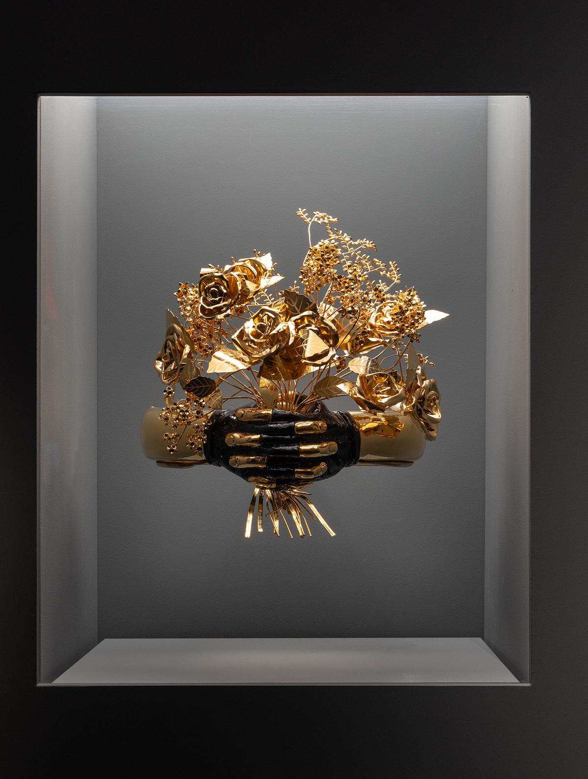 A golden bouquet of roses is held by a black skeletal hand inside a dimly lit shadow box, creating a stark, dramatic contrast and a surreal atmosphere.