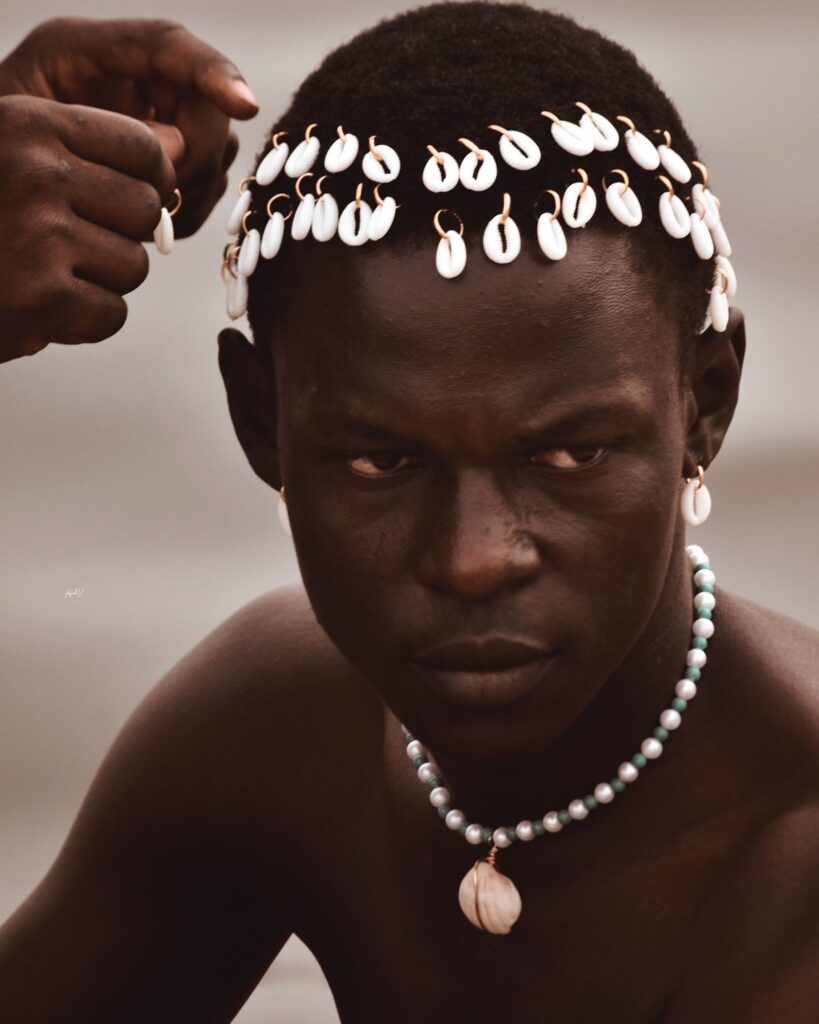 Close-up photograph of a young man wearing cowrie shells attached in rows across his hair, with matching shell earrings and a beaded necklace featuring a single cowrie pendant. Another hand adjusts the shells on his head.