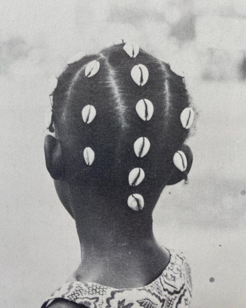 Black-and-white photograph showing the back of a young girl’s head, her hair neatly parted into rows and adorned with cowrie shells attached along each braid. She wears a patterned dress.