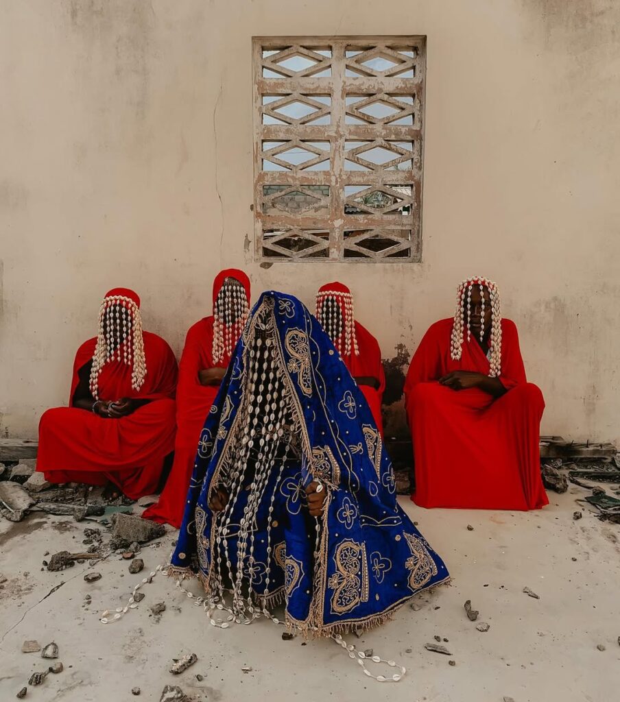Five figures sit against a wall, four dressed in red robes with veils of cowrie shells covering their faces, and one in the center draped in a deep blue, gold-embroidered cloak adorned with cascading cowrie shells. The shells hang in strands that veil the body, creating a powerful visual of ceremony and cultural symbolism.