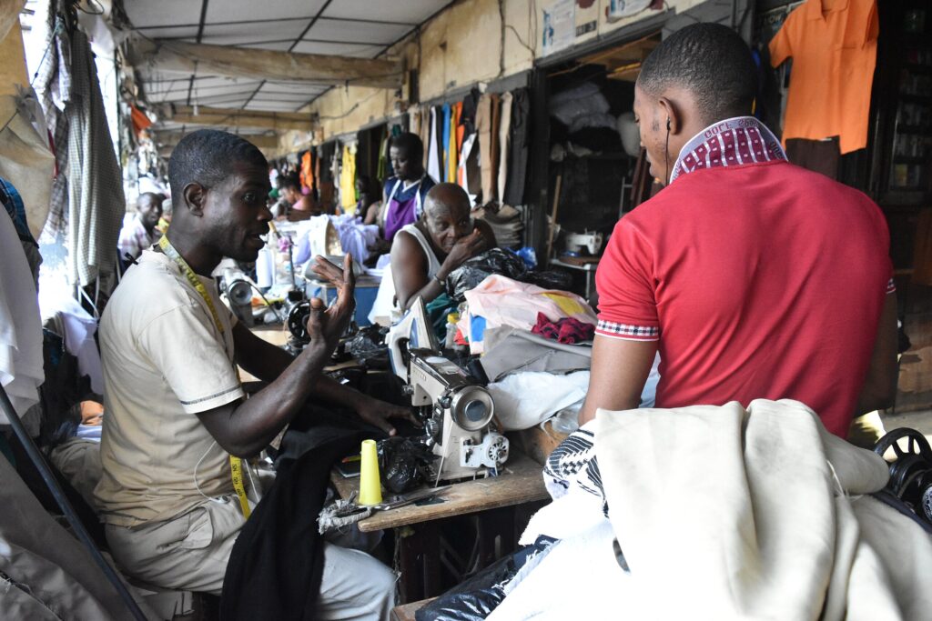 A group of tailors sewing and talking inside a busy market workshop, surrounded by fabrics and sewing machines.