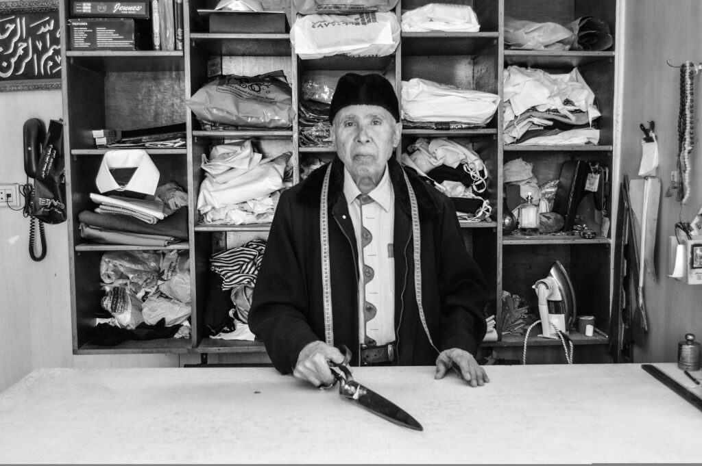 An elderly tailor in Egypt stands behind a worktable holding large scissors, shelves of fabric stacked behind him.
