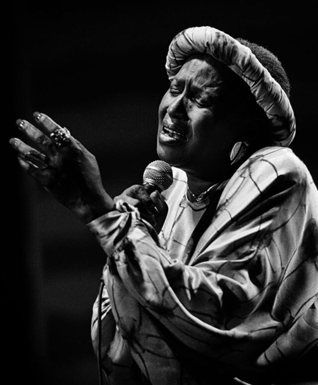 Black-and-white photograph of South African singer Miriam Makeba performing passionately with a microphone, wearing a patterned dress and headwrap.