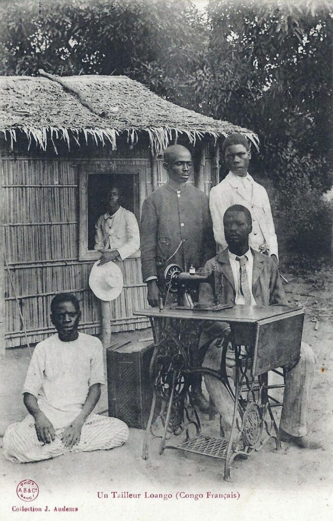A tailor in Loango, Congo Français, sits at a treadle sewing machine while men gather around him outside a bamboo hut, early 1900s.