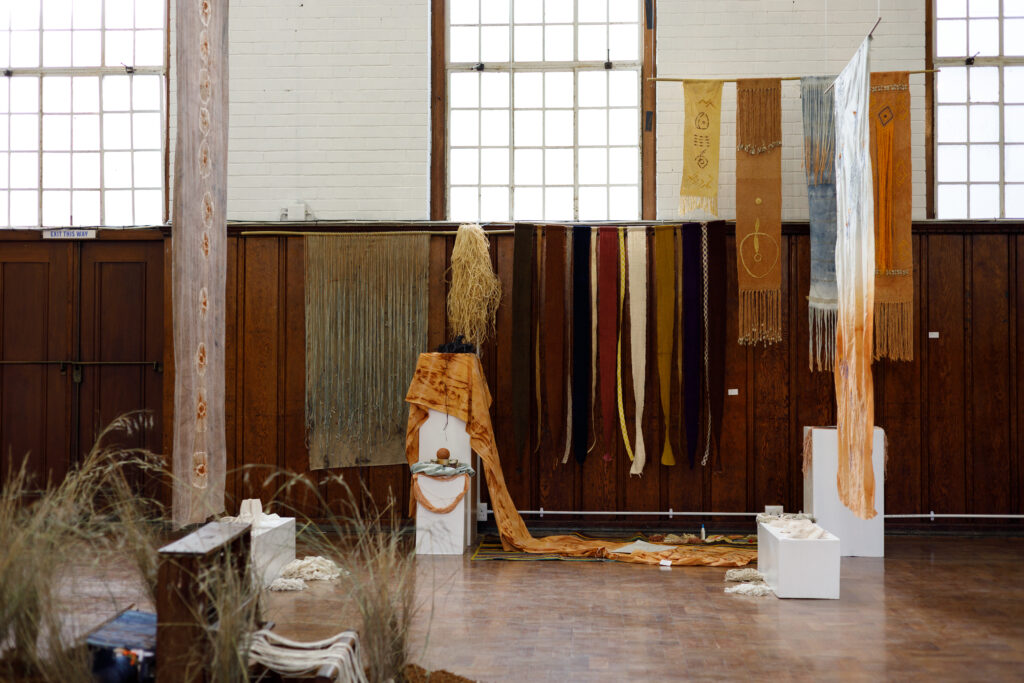 A textile installation featuring hanging woven pieces and hand-dyed fabrics in earthy tones of rust, beige, and indigo. Natural fibers and grasses are arranged on the floor and plinths, with long fabric strips cascading across the space, highlighting themes of visible mending and sustainable craft.