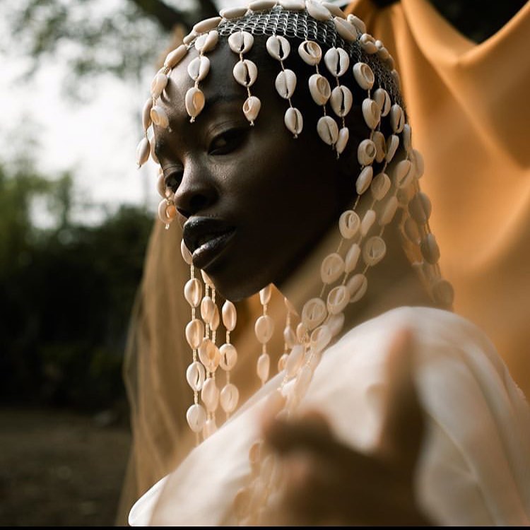 Portrait of a woman wearing a headpiece made of rows of cowrie shells that drape over her face and shoulders. She gazes into the camera with a commanding expression, dressed in flowing fabric, against a backdrop of golden cloth and soft natural light.