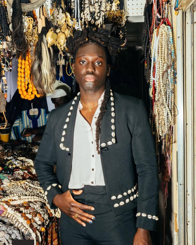 Portrait of a person standing in a market stall filled with jewelry and ornaments. They wear a black suit jacket and trousers embellished with cowrie shells along the collar, cuffs, and waistband, paired with a white shirt. Long locs frame their face as they look directly at the camera, surrounded by hanging beads, shells, and cultural artifacts.