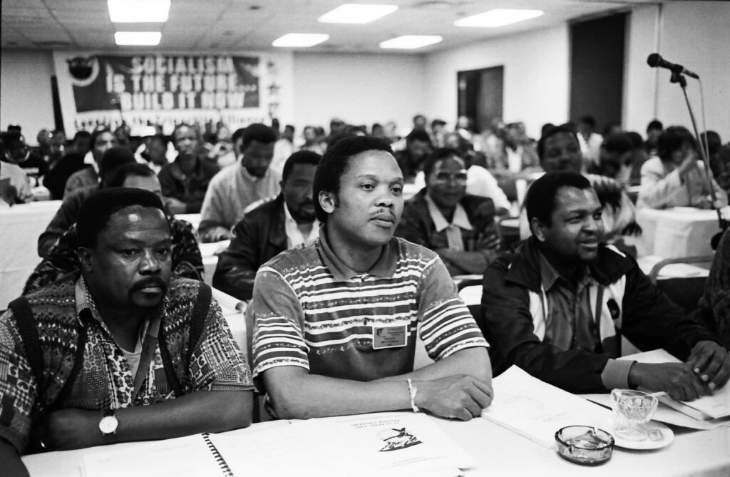 Black-and-white photograph of a crowded labour union meeting in South Africa during apartheid. Delegates sit with notebooks and papers; a banner hangs on the wall. Photograph © William Matlala.