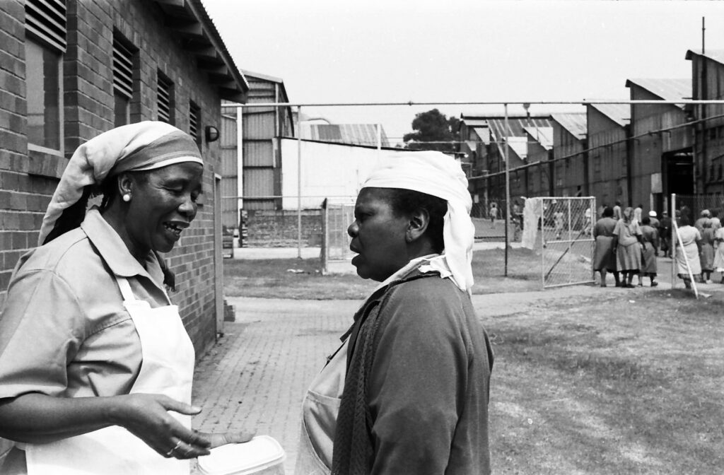 Black-and-white photo of two women standing and talking outside a factory building. One holds a container while others gather nearby.