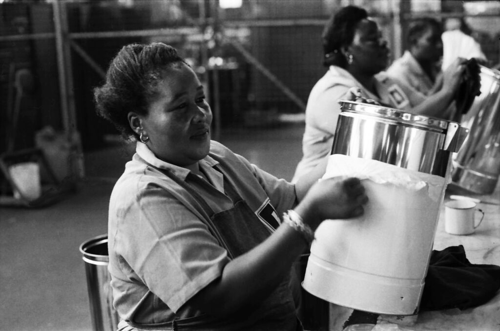 Black-and-white image of a woman polishing a steel container in a factory, South Africa. Photographed by William Matlala.
