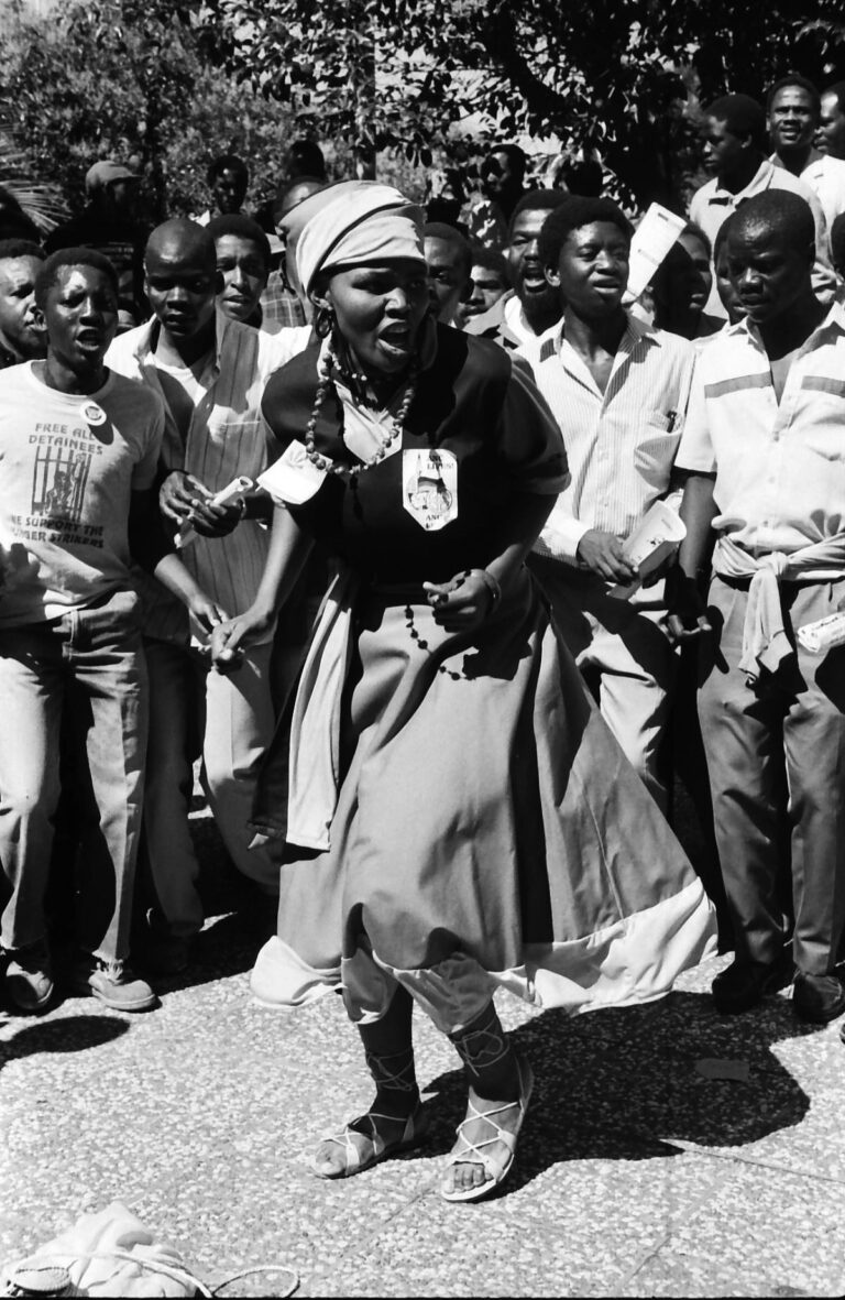 Black-and-white photograph of a woman in a headscarf and long skirt leading a protest, surrounded by men chanting behind her, captured by William Matlala in South Africa.