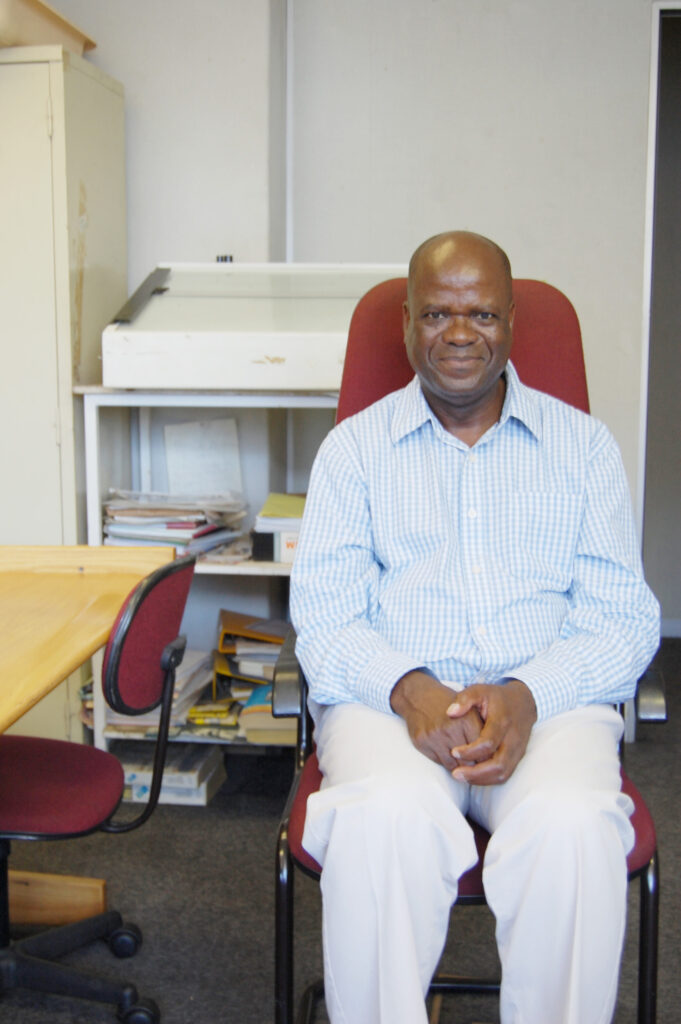 Colour portrait of William Matlala sitting in an office chair, wearing a checked shirt and white trousers, early 2000s.