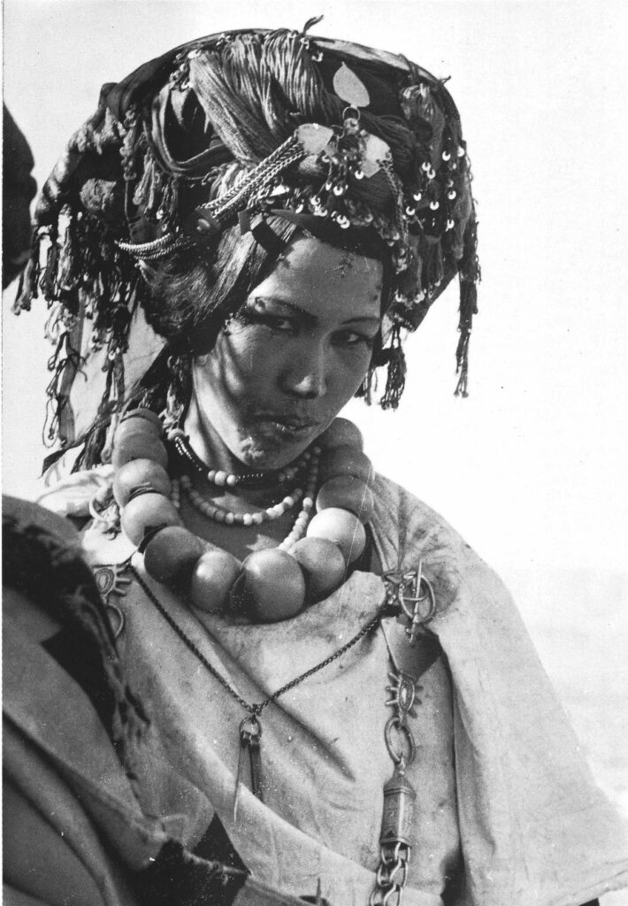 Black-and-white portrait of an Amazigh woman from Morocco’s Aït Seghrouchen tribe, wearing an elaborate headdress and large amber necklace.