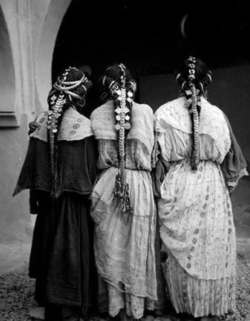 Black-and-white photograph of Amazigh women from Morocco, their long braids decorated with charms and beads, wearing traditional garments.