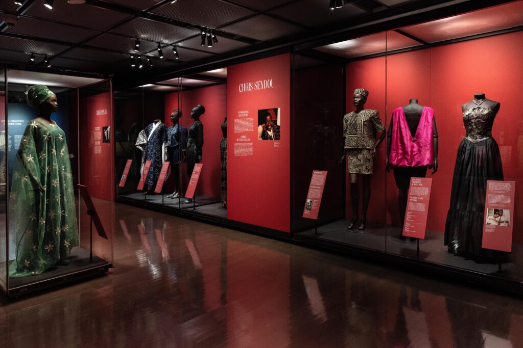 Africa Fashion Arrives in Montreal: A Global Conversation Continues 3 Series of mannequins dressed in wax-print and indigo pieces by Malian designer Chris Seydou, with red walls highlighting his biography.