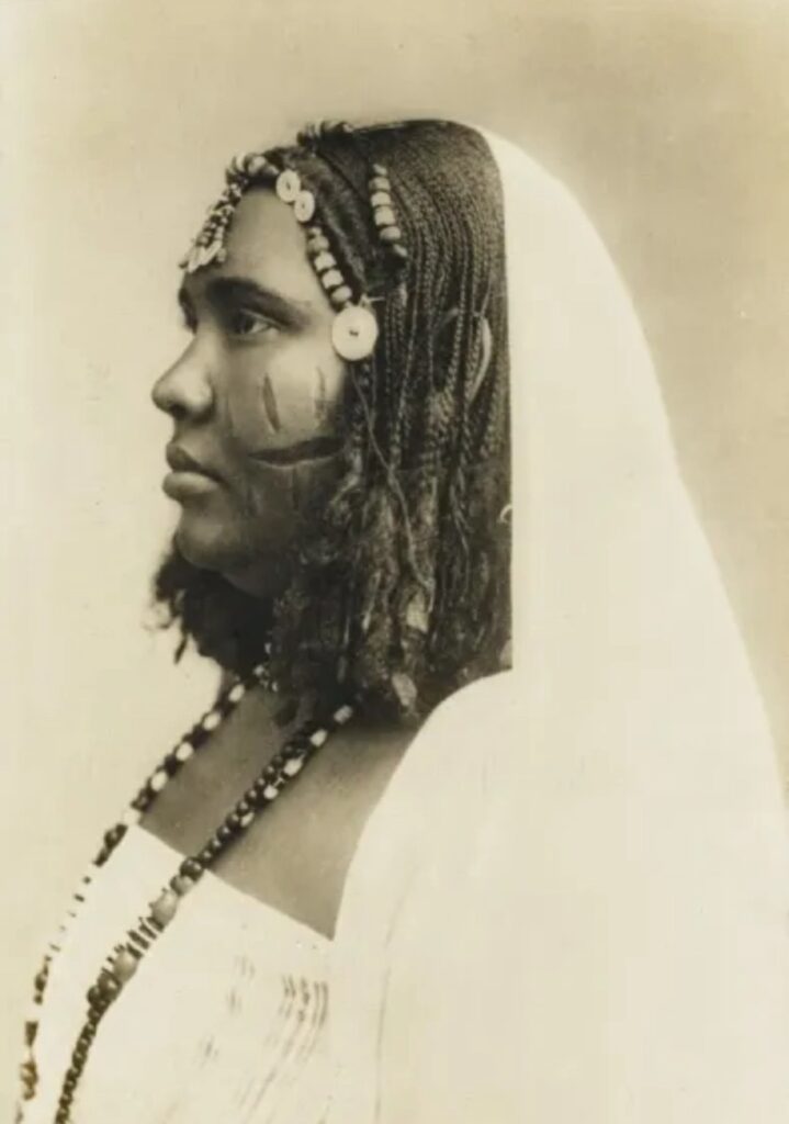 Archival portrait of a Sudanese woman with braided Mushat hairstyle and facial scarifications, wearing a light veil and bead necklace.