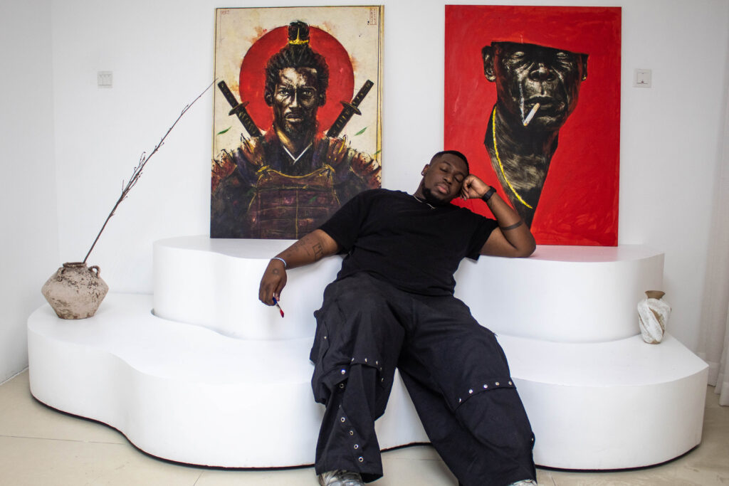 Anthony Azekwoh reclining on a white curved bench in his studio beneath two red-ground portraits