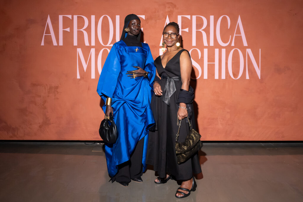 Two women stand before the wall reading Afrique / Africa Mode Fashion at the McCord Stewart Museum opening night. One wears a vivid cobalt blue draped ensemble with gold accessories, the other in an elegant black dress with statement earrings and a glittered handbag, capturing the evening’s blend of style and celebration.