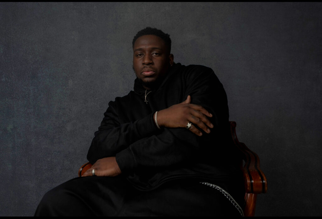 Portrait of the artist Anthony Azekwoh seated in black against a dark backdrop