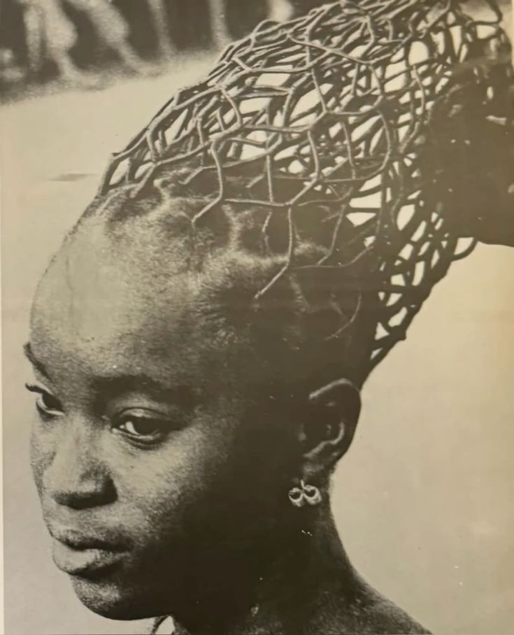 Black-and-white photograph of a Nigerian woman with an intricate braided lattice hairstyle extending upward, photographed by J.D. ’Okhai Ojeikere as part of his Hairstyles series, celebrating the sculptural artistry of Nigerian hair design.