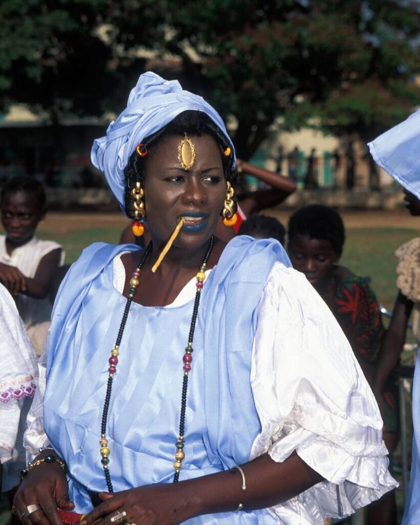 Wolof woman wearing a traditional blue boubou dress, ornate gold jewelry, and a blue moussor headwrap, photographed in Banjul, The Gambia, by Ariadne Van Zandbergen, depicting shared Senegambian cultural traditions.