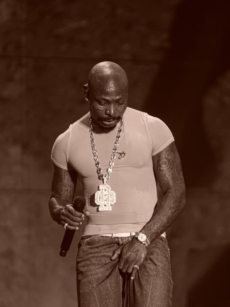 Portrait of Asake in a T-shirt and chain against a warm brown background