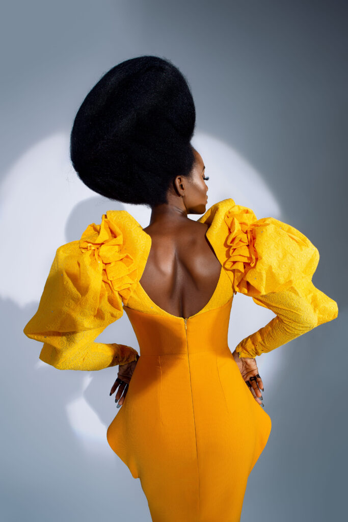 Back view of a model in a bright yellow gown with ruffled sleeves and a large afro, Umunwanyi Town Council collection