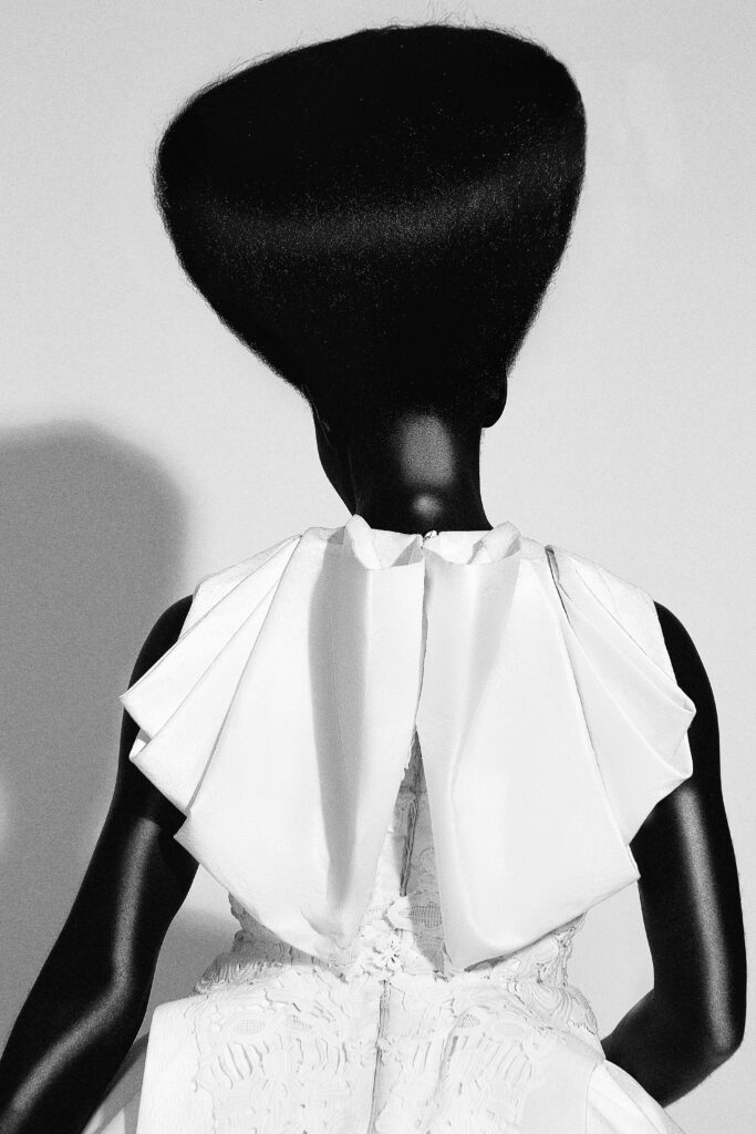 Model in a white structured top with sharply pointed shoulders and a tall sculpted afro, Umunwanyi Town Council collection