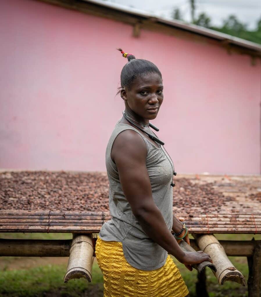 Portrait of a woman seated against a pink wall in a grey top and yellow wrapper, photographed by Nana Kofi Acquah