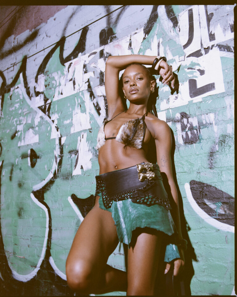 Model Lizette in PALMWINE IceCREAM SS26 — a hand-dyed leather skirt, studded belt with gold clasp, and feather-trimmed halter — photographed against a graffiti wall in New York by Myles Loftin.