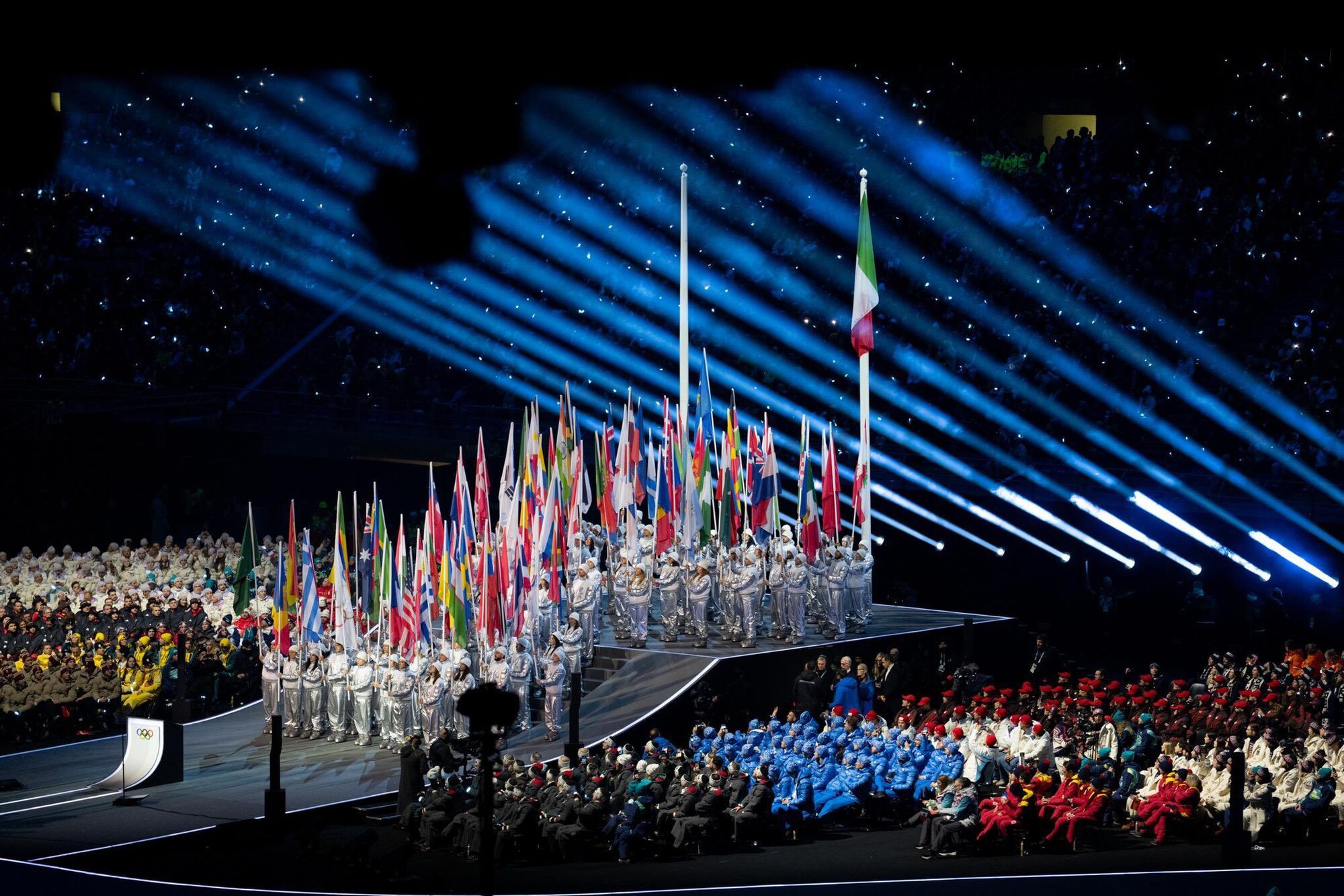 A wide shot of the illuminated stadium during the opening ceremony, featuring a vibrant display of international flags representing the global participation at the Milano Cortina 2026 Winter Olympics.