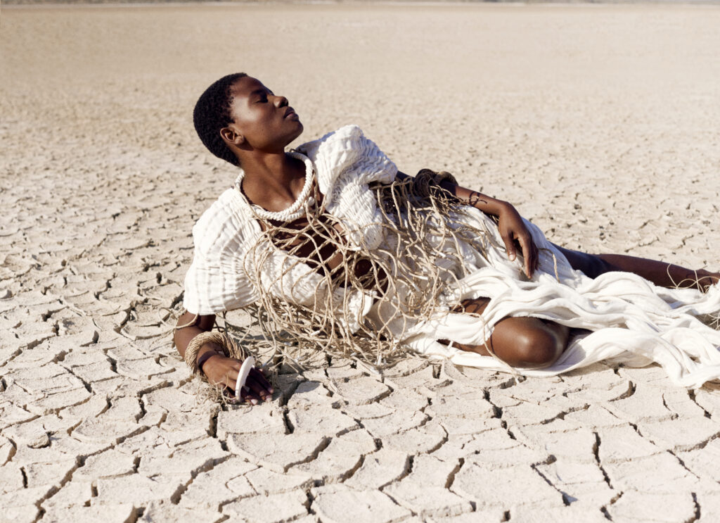 A fashion editorial photograph of a model reclining on a dry, cracked salt pan floor, wearing a textured, cream-colored garment from Ilkhe du Toit’s ‘Salt’ collection.