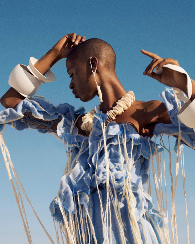 Ilkhe du Toit on Material-First Design and the Journey of Salt 3 A low-angle medium shot of a person with short hair wearing a blue and white patterned garment, posing with their hands near their face against a clear blue sky.