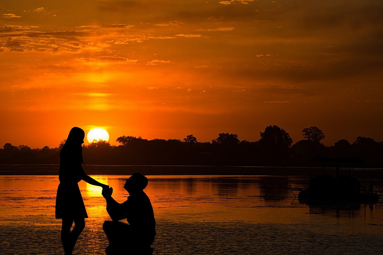 A romantic silhouette of a marriage proposal at sunset on an African riverbank, with vibrant orange skies reflecting on the water and trees in the distance.
